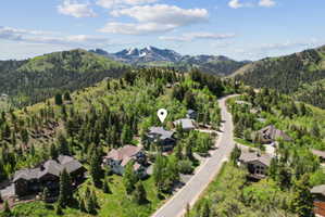 Mountainous terrain featuring a paved road, scattered residences, and extensive tree coverage under a partly cloudy sky