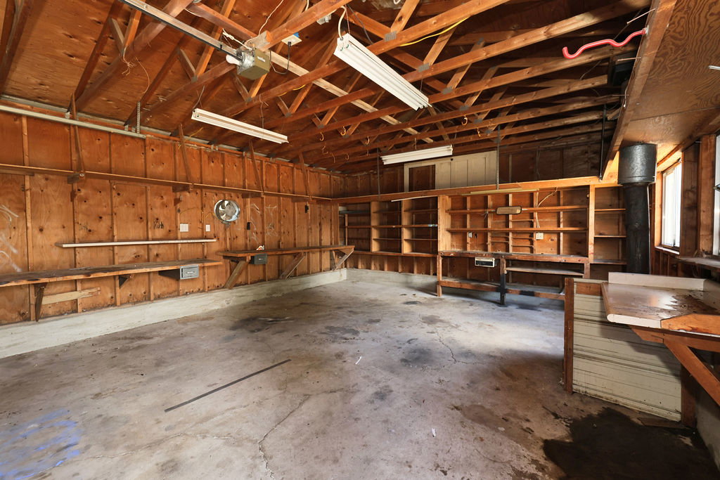 Spacious garage featuring exposed wood beam ceiling, concrete flooring, extensive built-in shelving, multiple work surfaces, and overhead fluorescent lighting