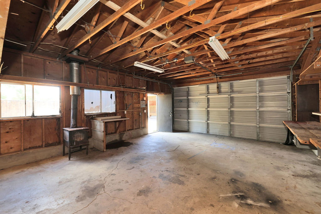 Spacious garage featuring exposed wood beam ceilings, concrete flooring, a roll-up garage door, multiple windows, and a built-in wood-burning stove with flue