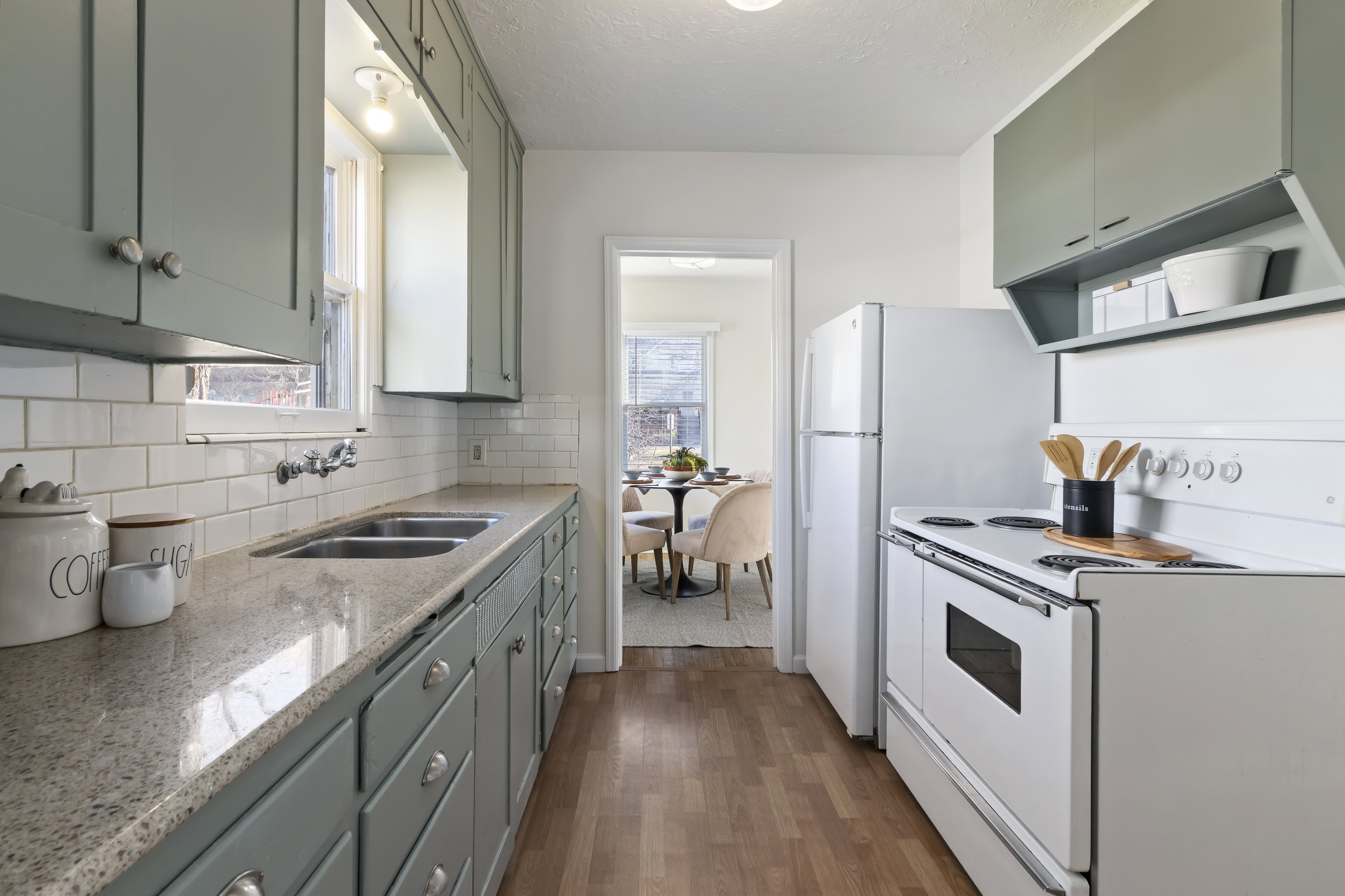 Kitchen featuring light green cabinetry, a double basin stainless steel sink, a subway tile backsplash, light-toned countertops, and wood-finish flooring