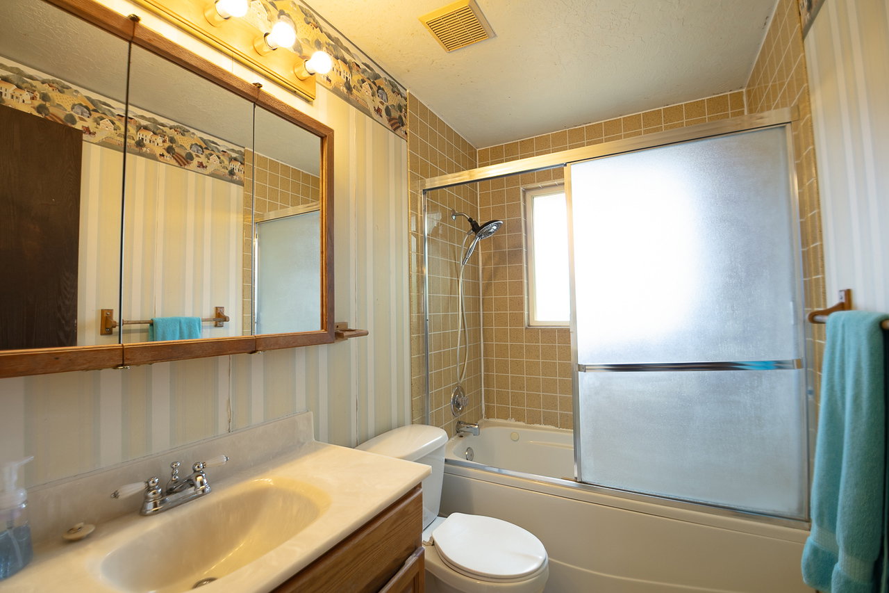 Bathroom featuring a built-in tub with a sliding glass enclosure, light-toned square tile surround, and a window
