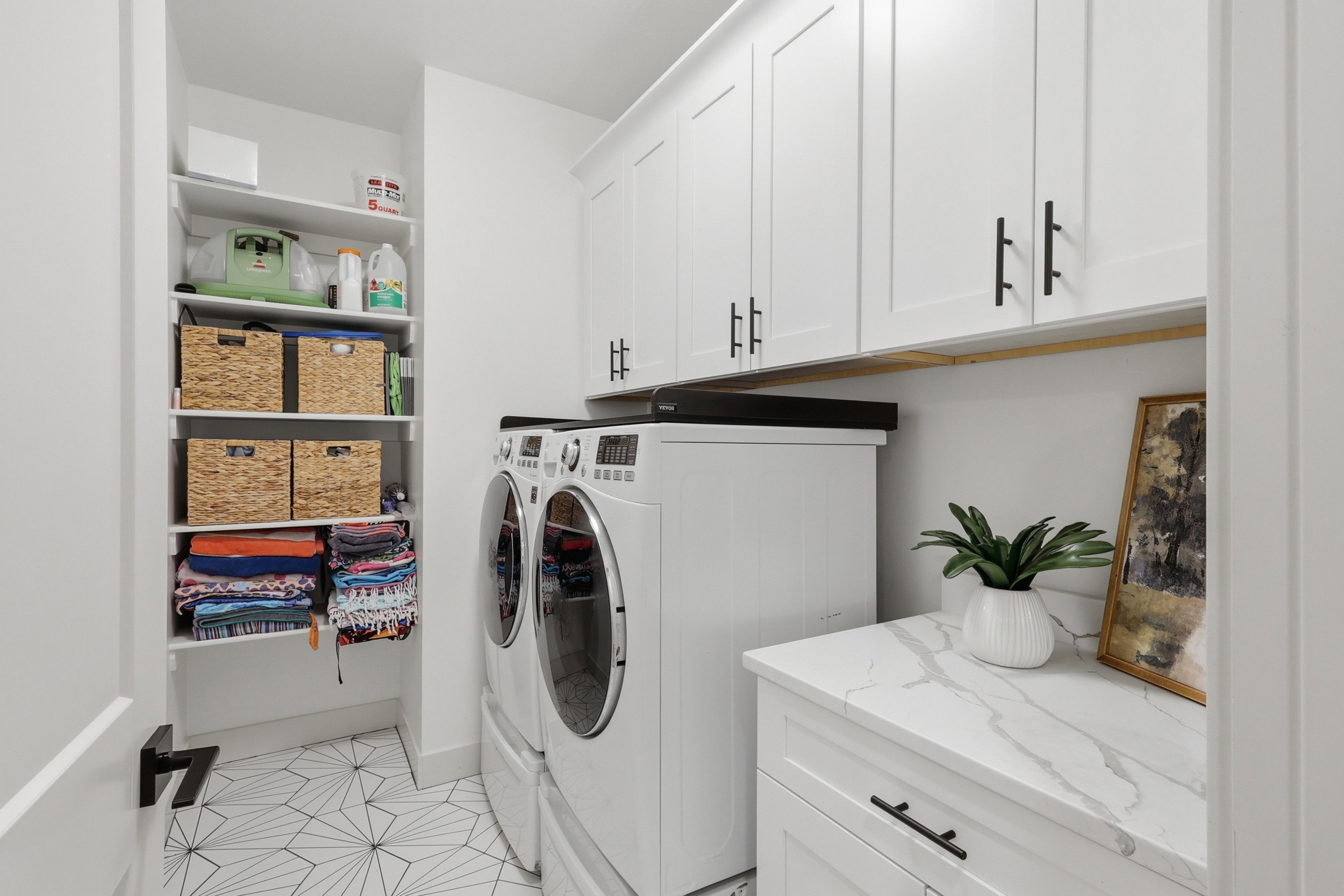 Dedicated laundry space featuring stacked appliances, overhead white shaker cabinetry with black hardware, an integrated counter with a marble-finish surface, and a built-in shelving unit