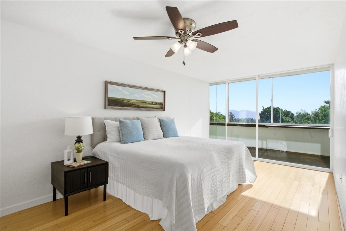 Bright room featuring expansive sliding glass doors, a private balcony, wood-finish flooring, a ceiling fan with integrated lighting, and a light-colored headboard
