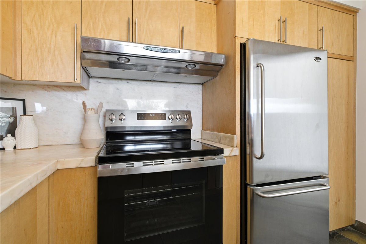 Kitchen area featuring light wood cabinetry, stainless steel appliances including a range with an overhead hood, and a refrigerator