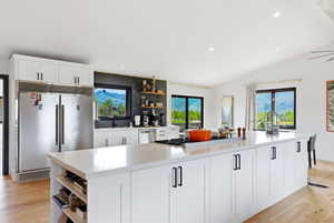 Spacious kitchen featuring a large white island with integrated cooktop, light wood-finish flooring, white cabinetry with black hardware, recessed lighting, and large windows providing natural light