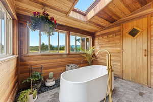 Freestanding soaking tub with a brass floor-mounted faucet, surrounded by wood-paneled walls and ceiling, a skylight, and large windows revealing a mountain backdrop