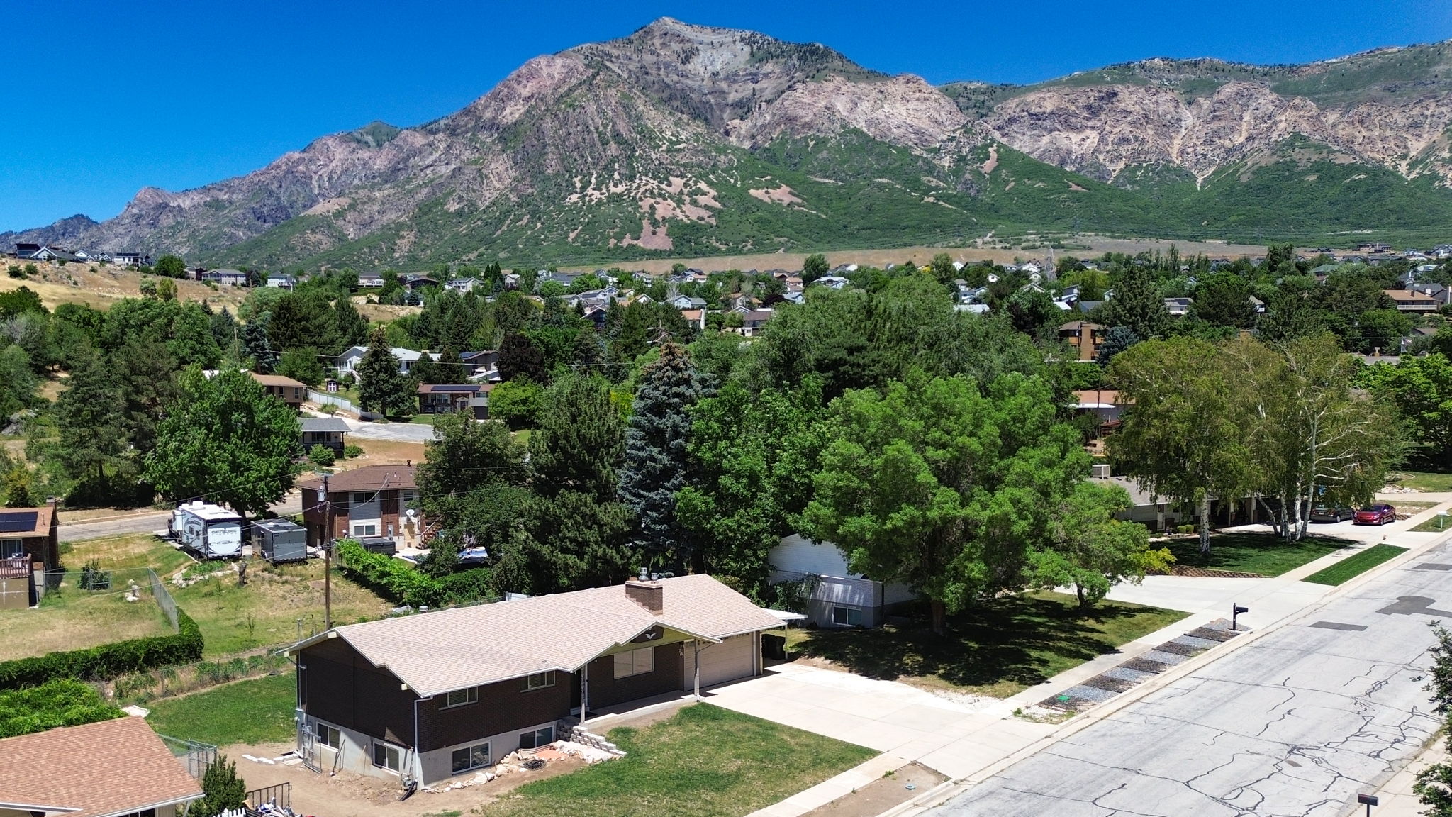 Mountain backdrop showcasing a residential property with a brown-roofed structure, attached carport, concrete driveway, and established landscaping