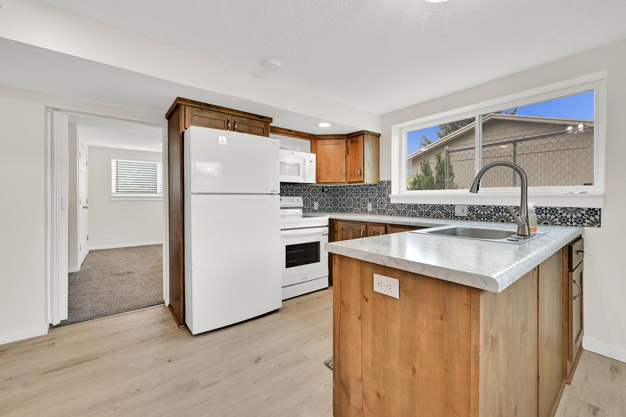 Kitchen featuring wood-finish flooring, a patterned tile backsplash, wood cabinetry, an island with a built-in sink, and a white appliance suite