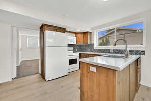 Kitchen featuring wood-finish flooring, a patterned tile backsplash, wood cabinetry, an island with a built-in sink, and a white appliance suite