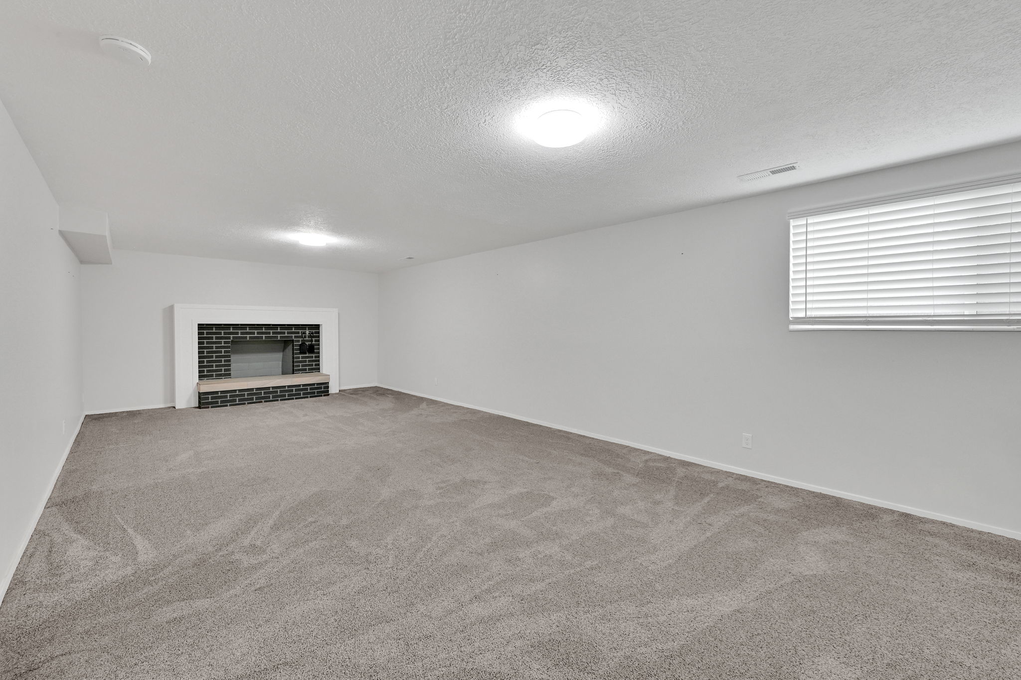 Spacious room with a brick-clad fireplace and hearth, neutral carpet, white walls, and a window with blinds