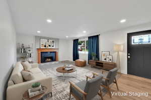Living area featuring a fireplace with a wood mantle and tile surround, recessed lighting, wood-finish flooring, a contemporary dark entry door with a transom window, and windows with dark-colored curtains. Room was virtually staged.
