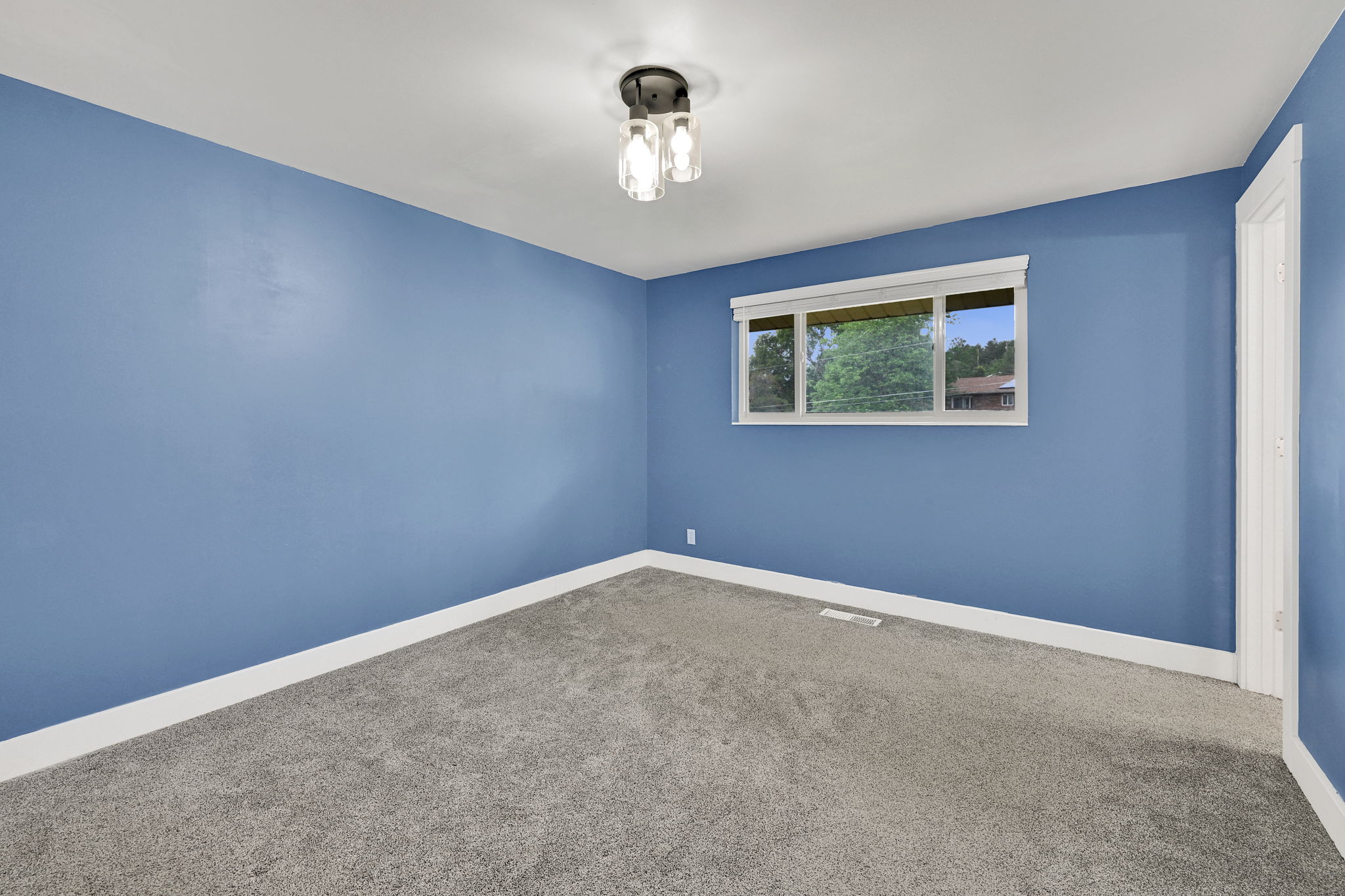 Spacious room with blue walls and light gray carpeting, featuring a multi-pane window with white trim, a modern three-light ceiling fixture, and white baseboards