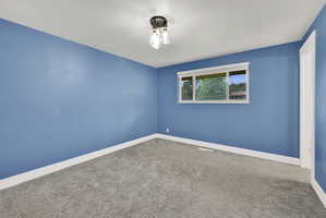 Spacious room with blue walls and light gray carpeting, featuring a multi-pane window with white trim, a modern three-light ceiling fixture, and white baseboards