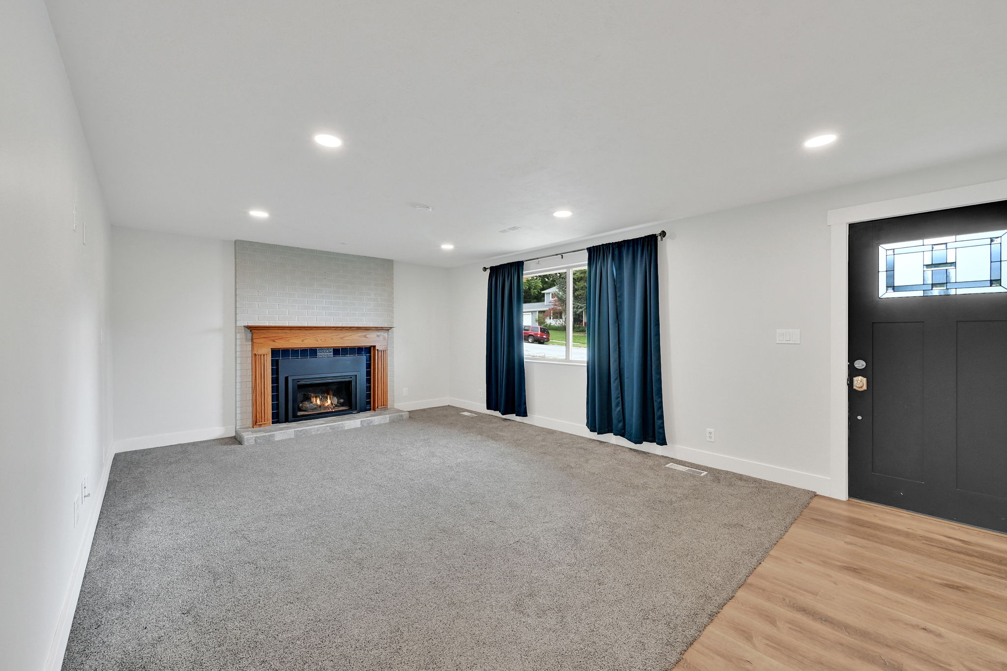 Neutral interior featuring a fireplace with a light brick surround and wood-finish mantel, recessed lighting, wood-finish flooring at the entry, and a dark paneled door with decorative glass