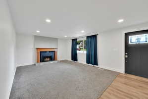 Neutral interior featuring a fireplace with a light brick surround and wood-finish mantel, recessed lighting, wood-finish flooring at the entry, and a dark paneled door with decorative glass