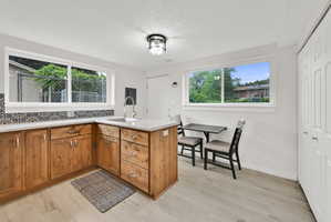 Light-filled kitchen featuring natural wood cabinetry, light-toned countertops, a patterned tile backsplash, wood-finish flooring, and a stainless steel sink with a high-arc faucet