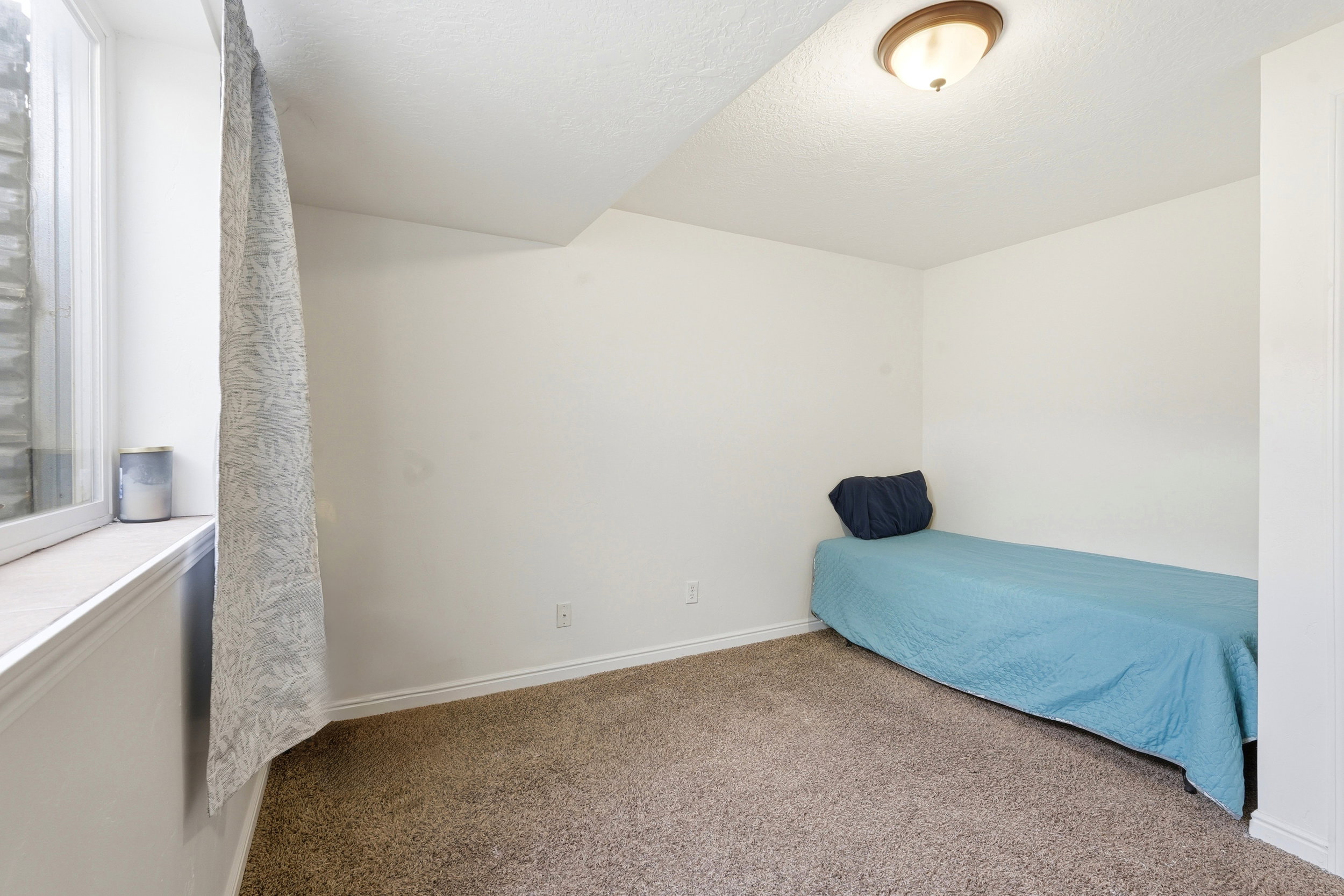 Carpeted room with a ceiling-mounted light fixture, white walls, and a window featuring a patterned curtain