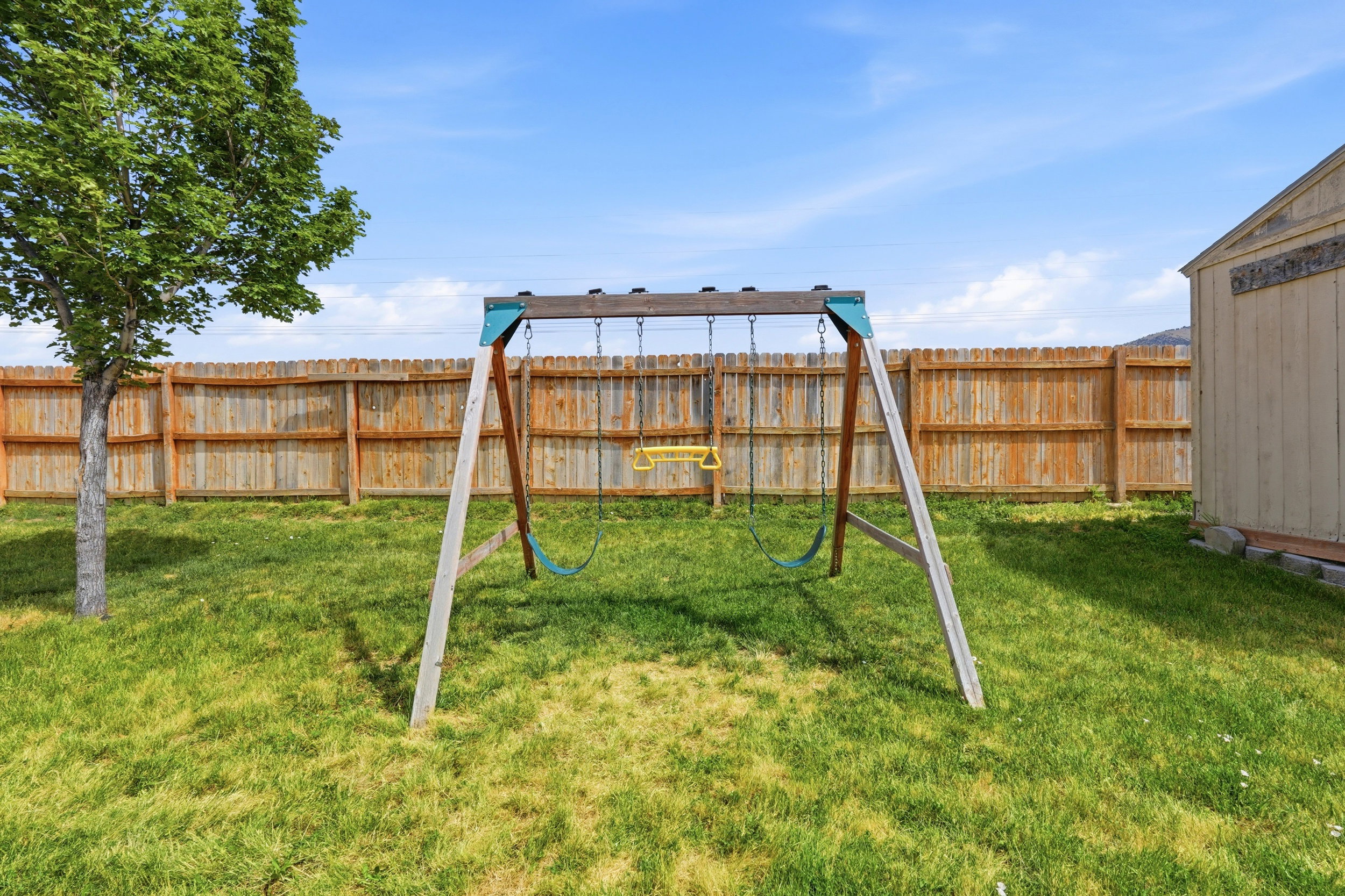 Expansive green lawn featuring a wooden swing set, a mature tree, and a light-colored storage shed