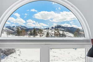 Arched window framing a mountain landscape with snow-covered peaks, pine trees, and a clear sky with scattered clouds