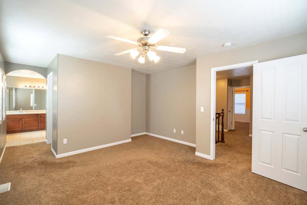 Carpeted room with a ceiling fan, featuring a wide arched doorway leading to a bathroom with a dual vanity, large mirror, and tiled flooring