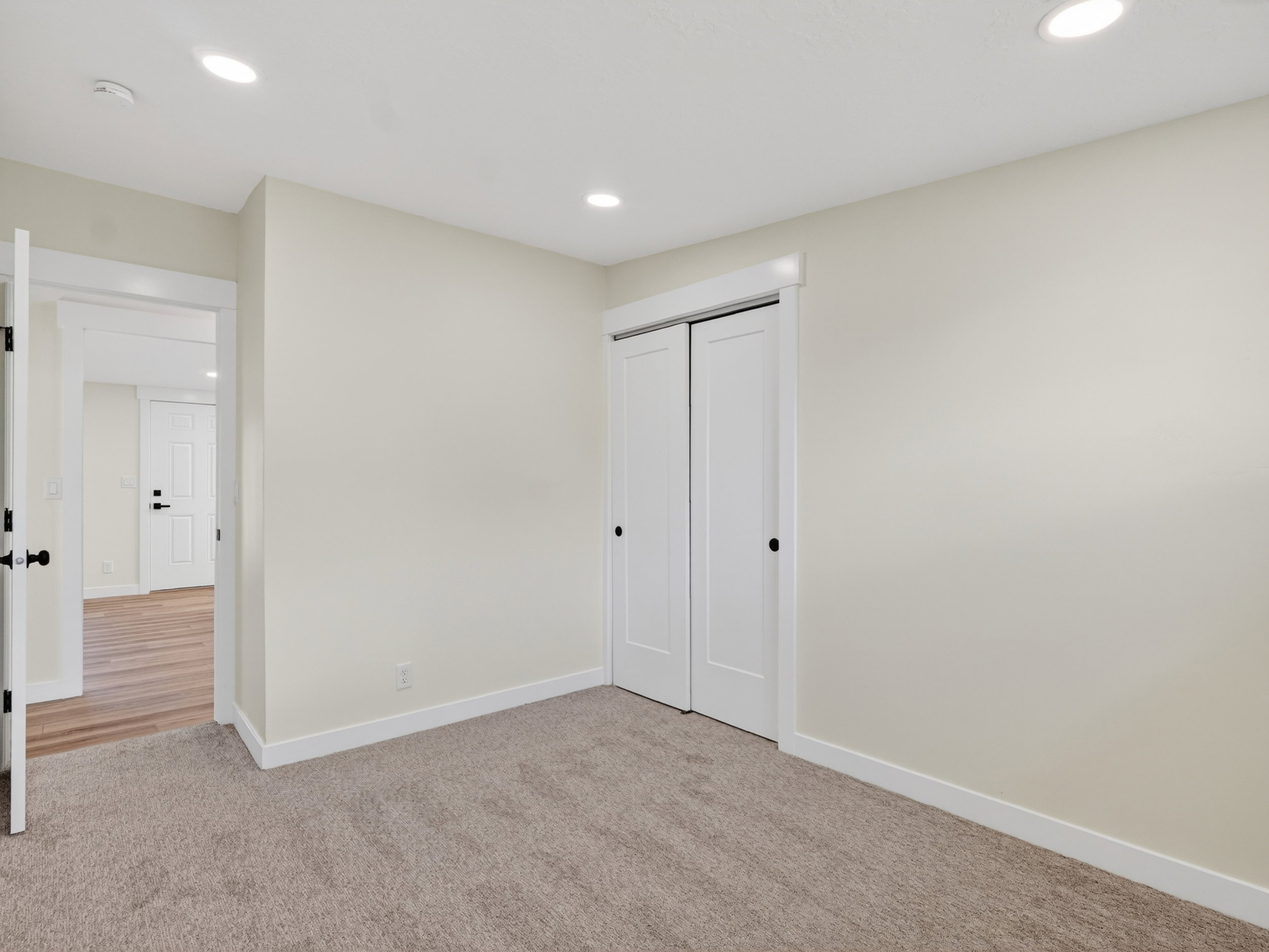 Neutral tone carpeting, recessed ceiling lighting, a pair of bi-fold closet doors with matte black hardware, and crisp white baseboards
