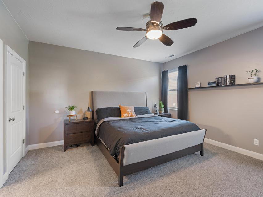 Carpeted bedroom featuring a ceiling fan with integrated lighting, a built-in wall shelf, a single window with dark drapes, and a white paneled door with a brass knob