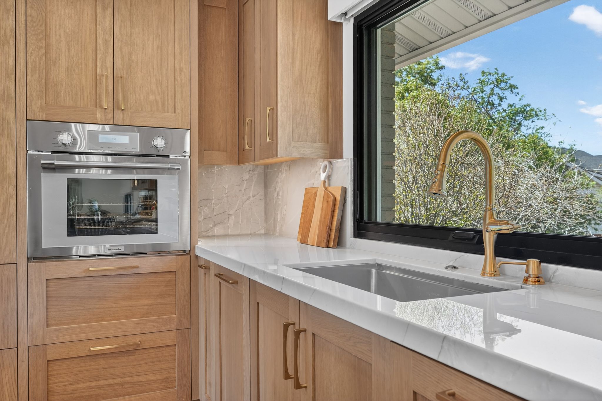 Custom cabinetry in a light wood finish, integrated stainless steel wall oven,  natural stone countertops with subtle veining, undermount sink, and a gold-tone high-arc faucet