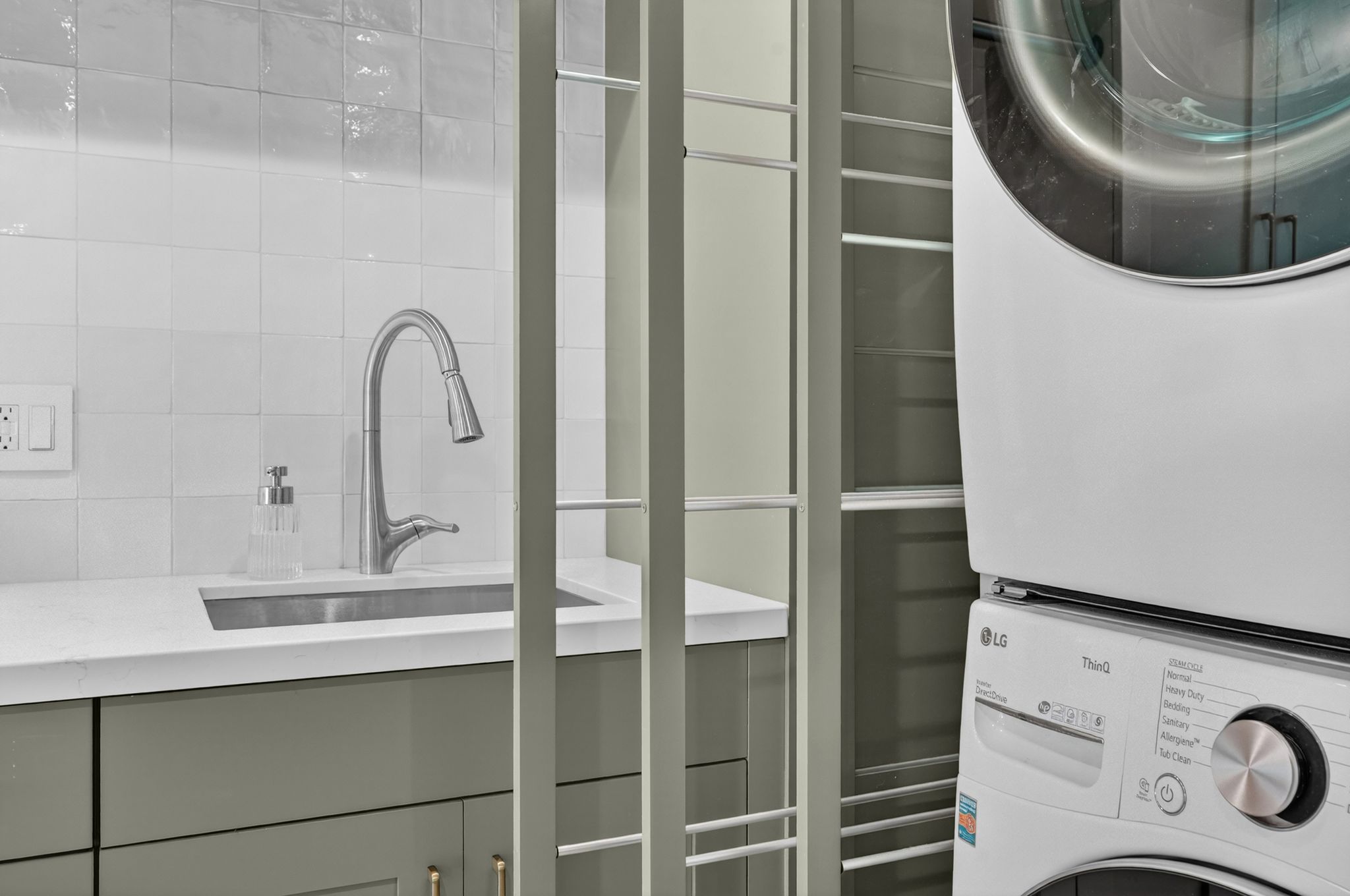 Dedicated laundry area featuring a stacked washer and dryer, a utility sink with a gooseneck faucet, light-colored cabinetry, and a white tiled backsplash