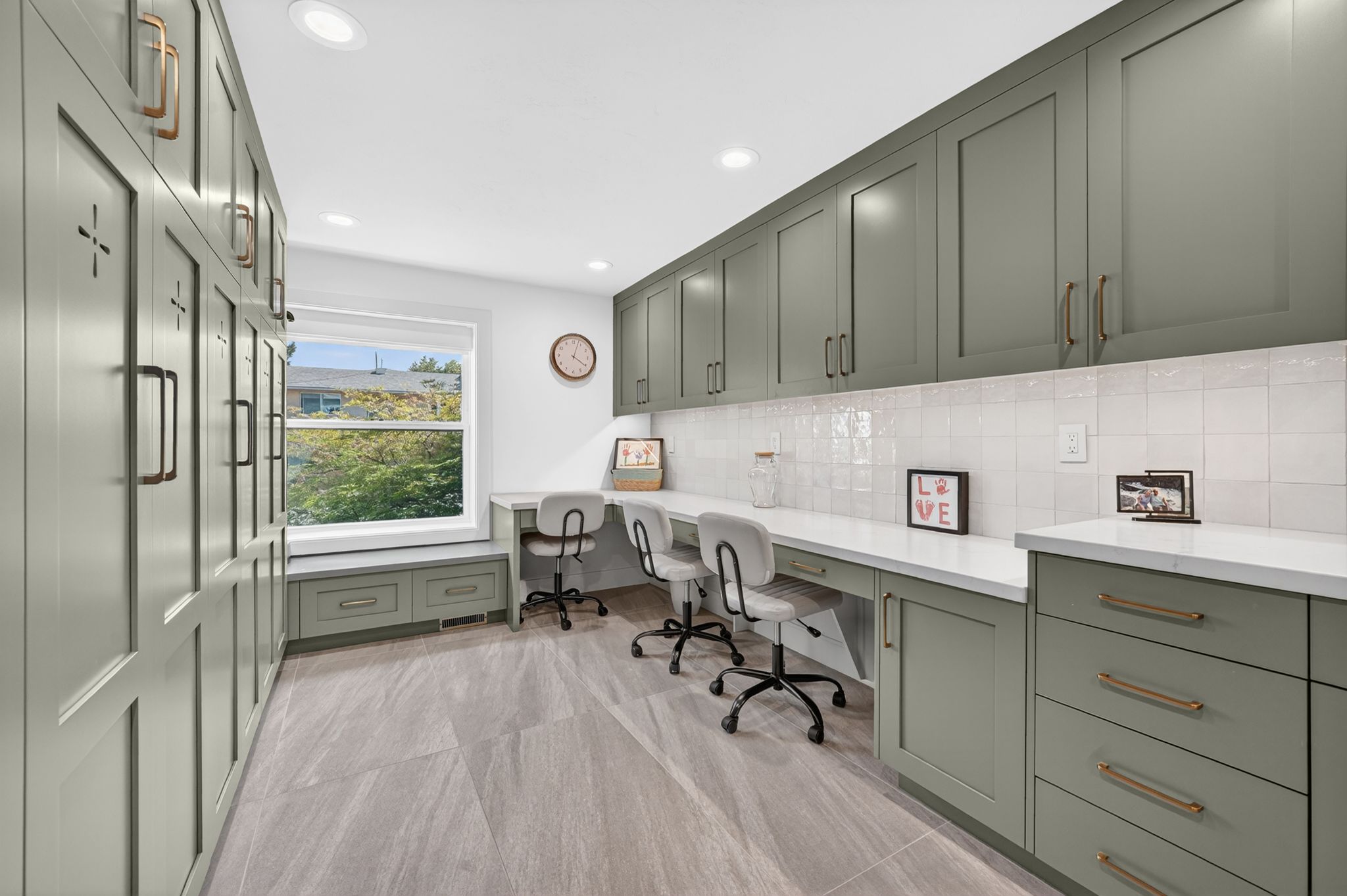 Dedicated workspace featuring extensive sage green cabinetry with bronze hardware, a built-in desk area with dual workstations, stone countertops, and a white subway tile backsplash