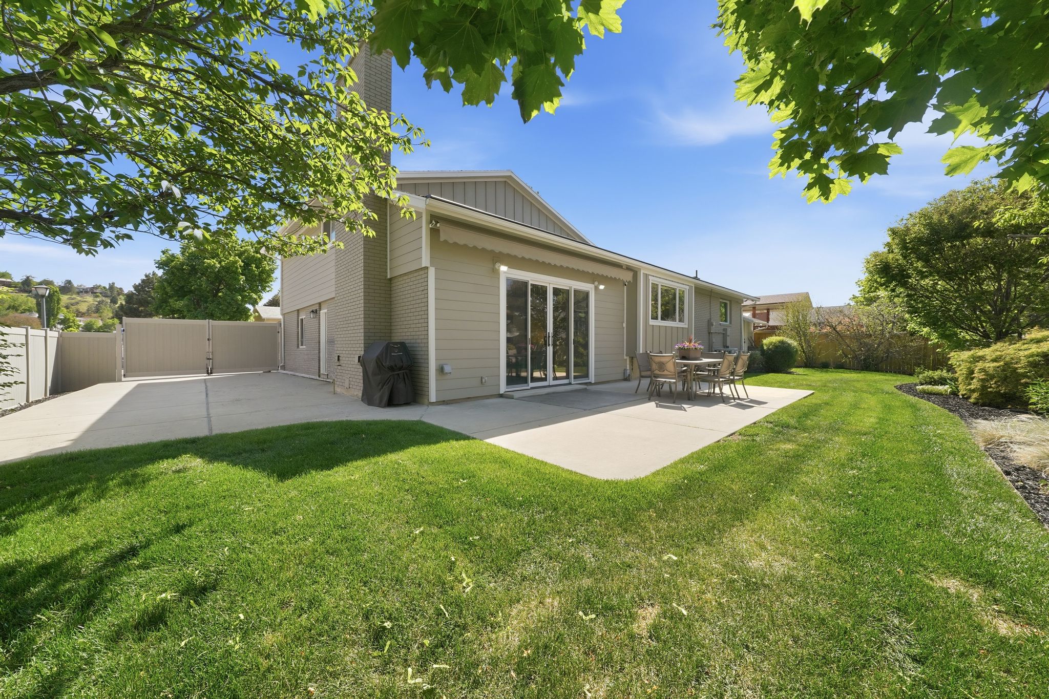 Expansive rear yard featuring a large concrete patio with retracting awning, manicured lawn, mature trees, and a concrete RV parking