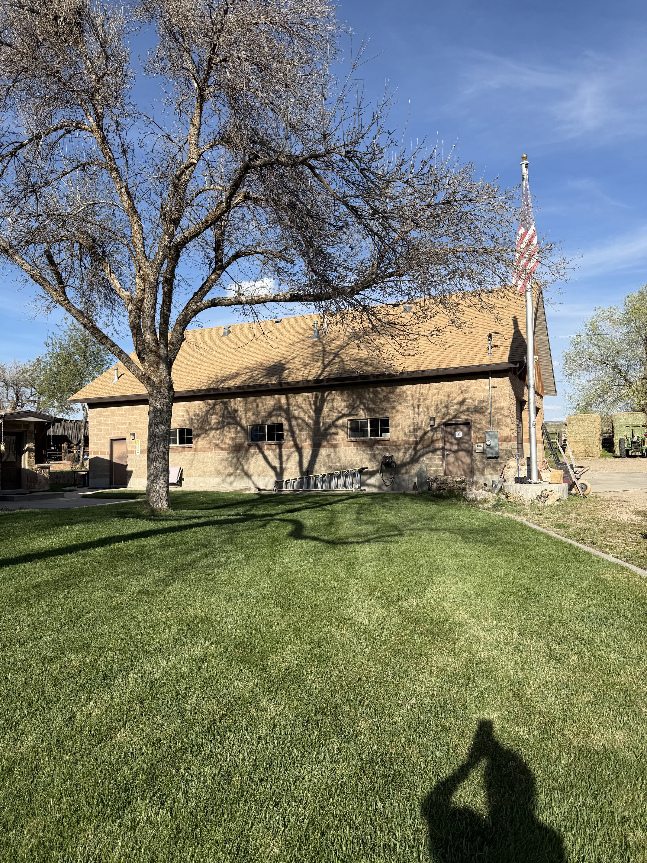 Expansive green lawn with mature tree, brick exterior structure, brown shingle roof, multiple windows, and a flagpole