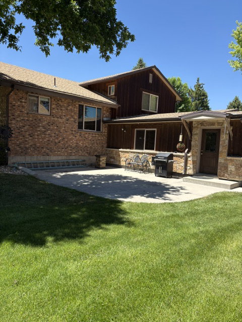 Expansive backyard featuring a lush lawn, a concrete patio, brick and wood-paneled exterior, a stone-clad entry, and multiple exterior windows