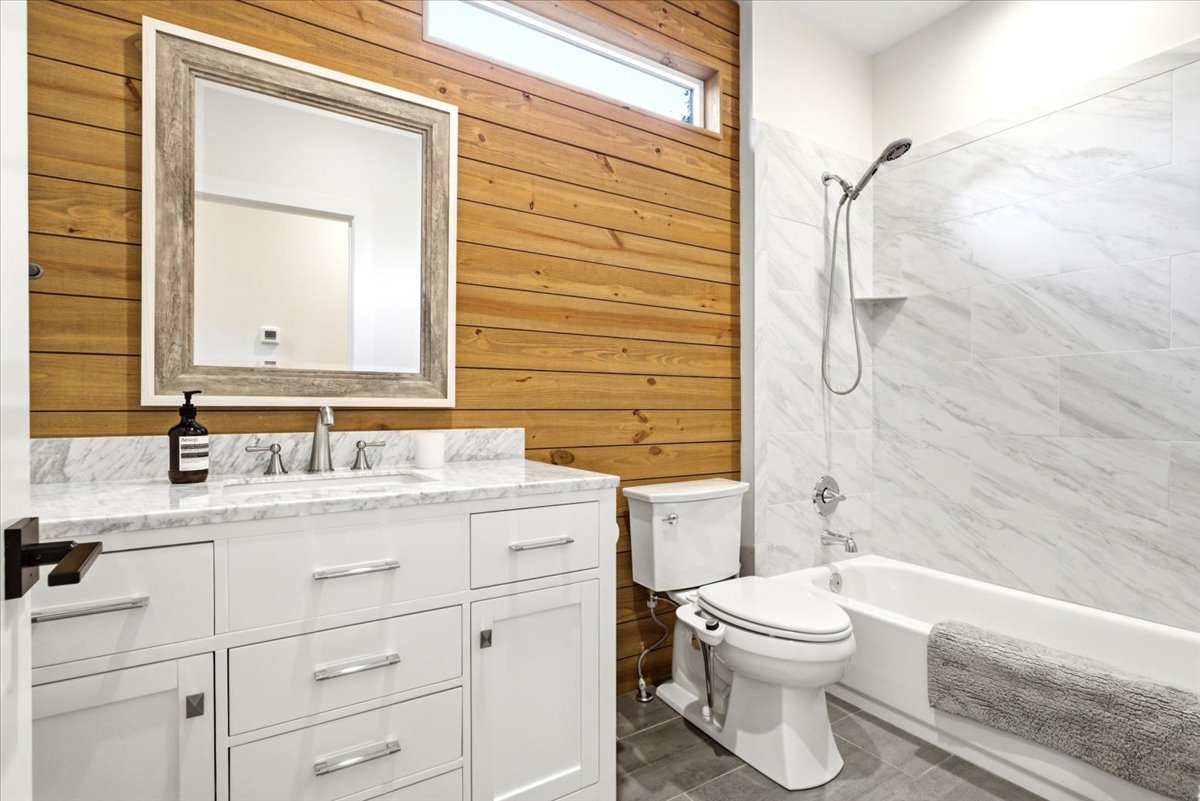 Bathroom featuring a white vanity with a marble countertop and undermount sink, wood-paneled accent wall, a framed mirror, a tub/shower combination with marble-look tile surround, and grey floor tiles