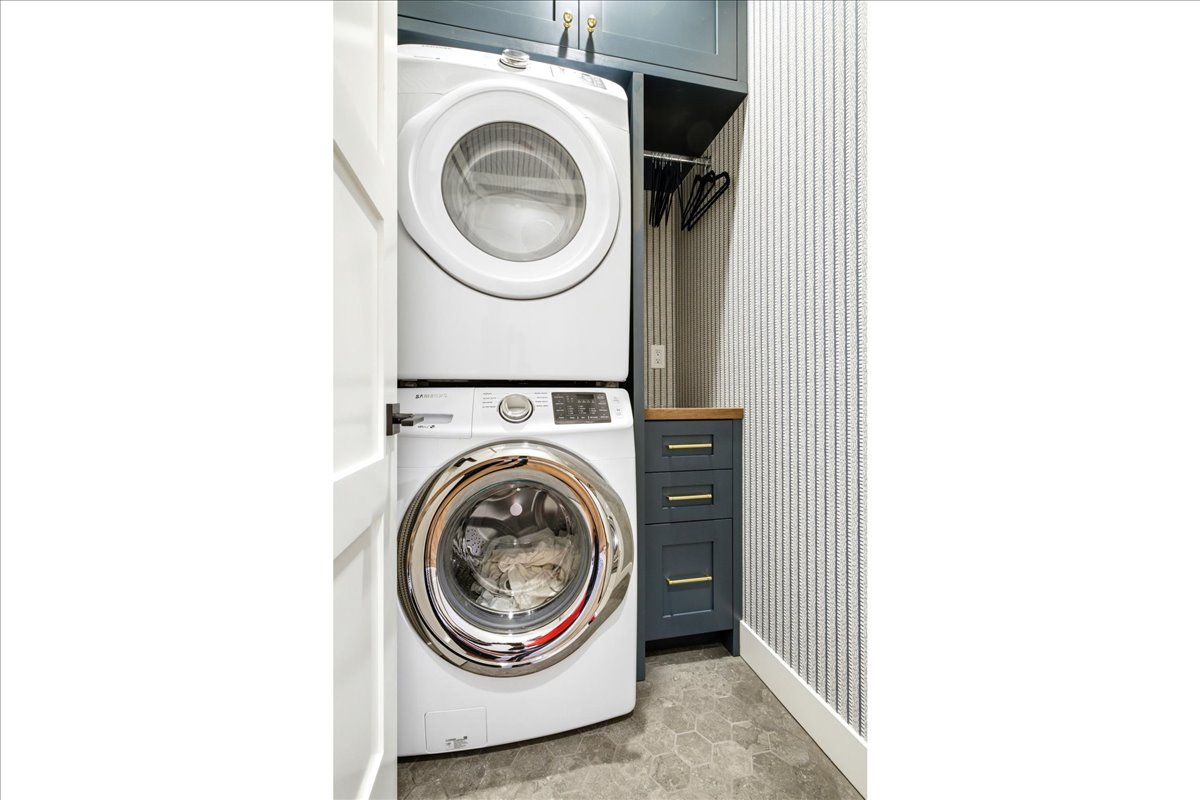 Laundry area featuring a stacked washer and dryer, custom cabinetry with a wood-finish countertop, hexagonal floor tiles, and striped wallpaper