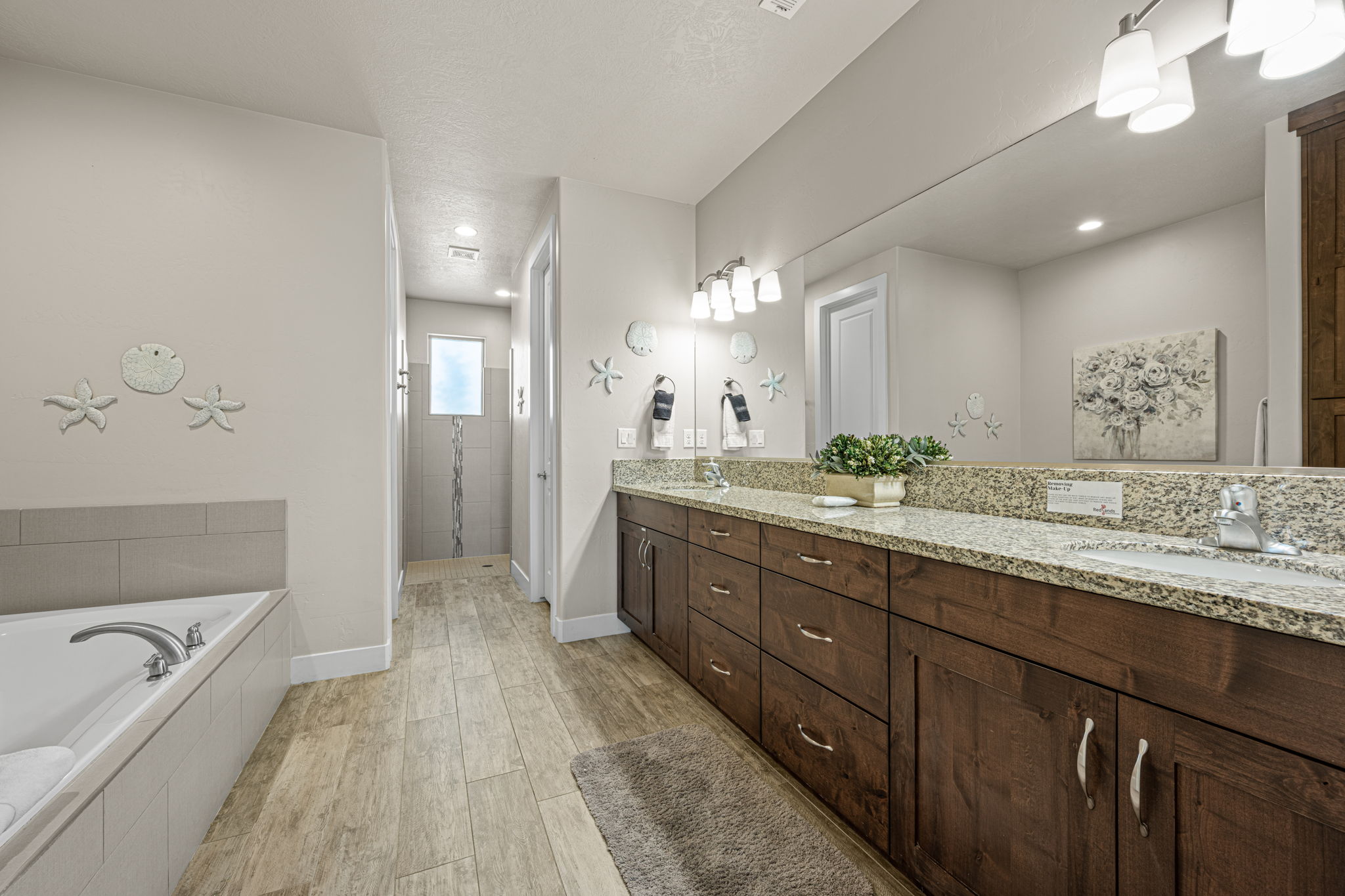 Spacious bathroom featuring a dual vanity with a light-toned countertop, extensive under-sink storage, a built-in soaking tub, a separate shower stall with tile surround, and wood-finish flooring
