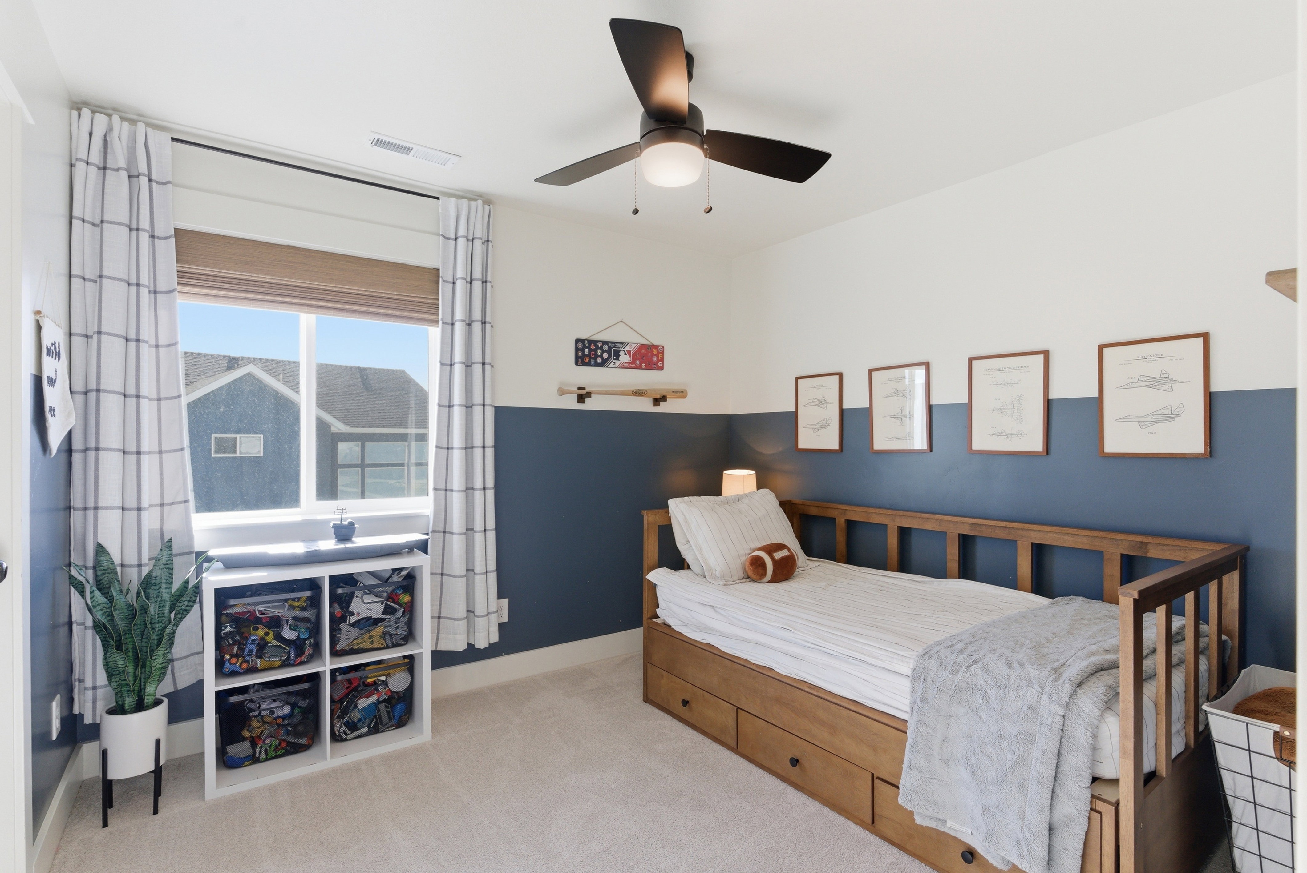 Light-filled bedroom featuring a large window with blinds and plaid curtains, a ceiling fan with integrated lighting, two-tone painted walls, and neutral carpet