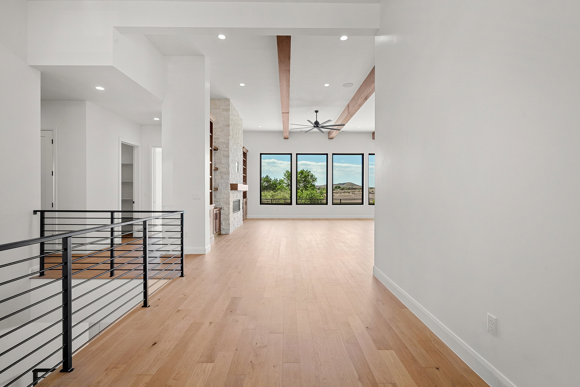 Expansive room featuring light wood-finish flooring, a stone-clad fireplace with built-in shelving, triple picture windows, exposed ceiling beams, and recessed lighting
