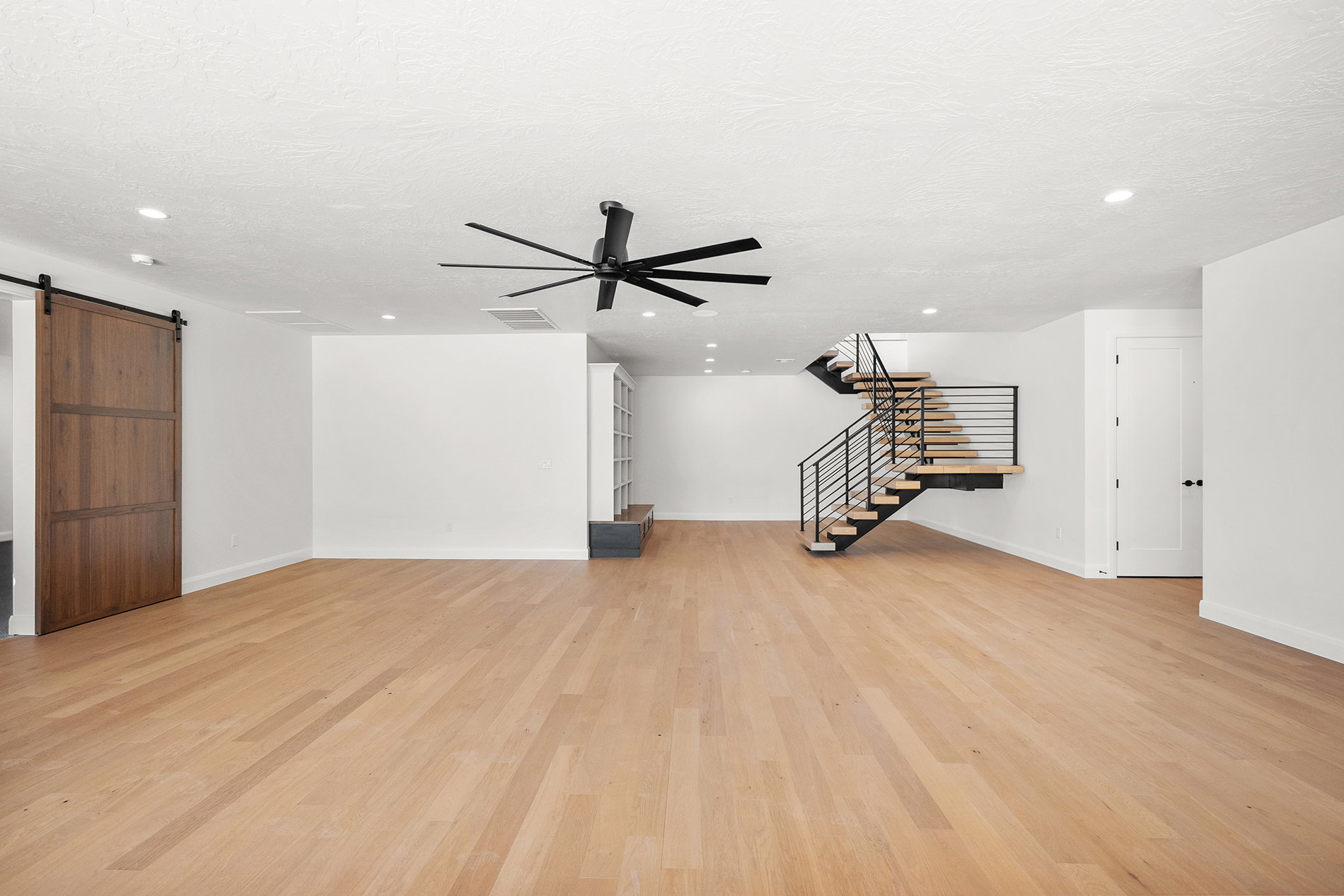 Spacious living area featuring wood-finish flooring, a modern floating staircase with metal railings, a large ceiling fan, recessed lighting, and a wood-panel barn door