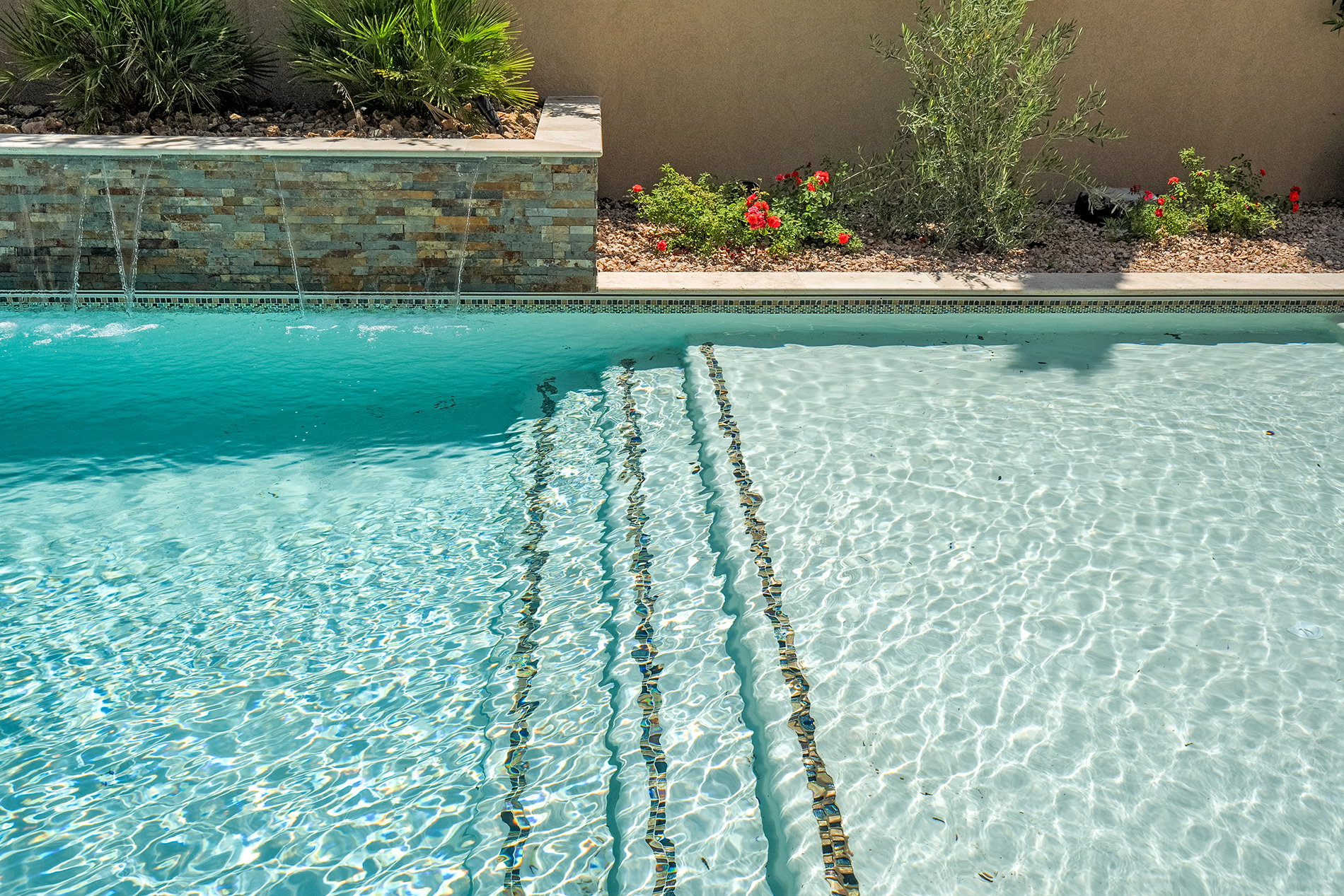 Swimming pool featuring a stacked stone water feature, integrated entry steps with mosaic tile accents, and a surrounding planter with desert landscaping