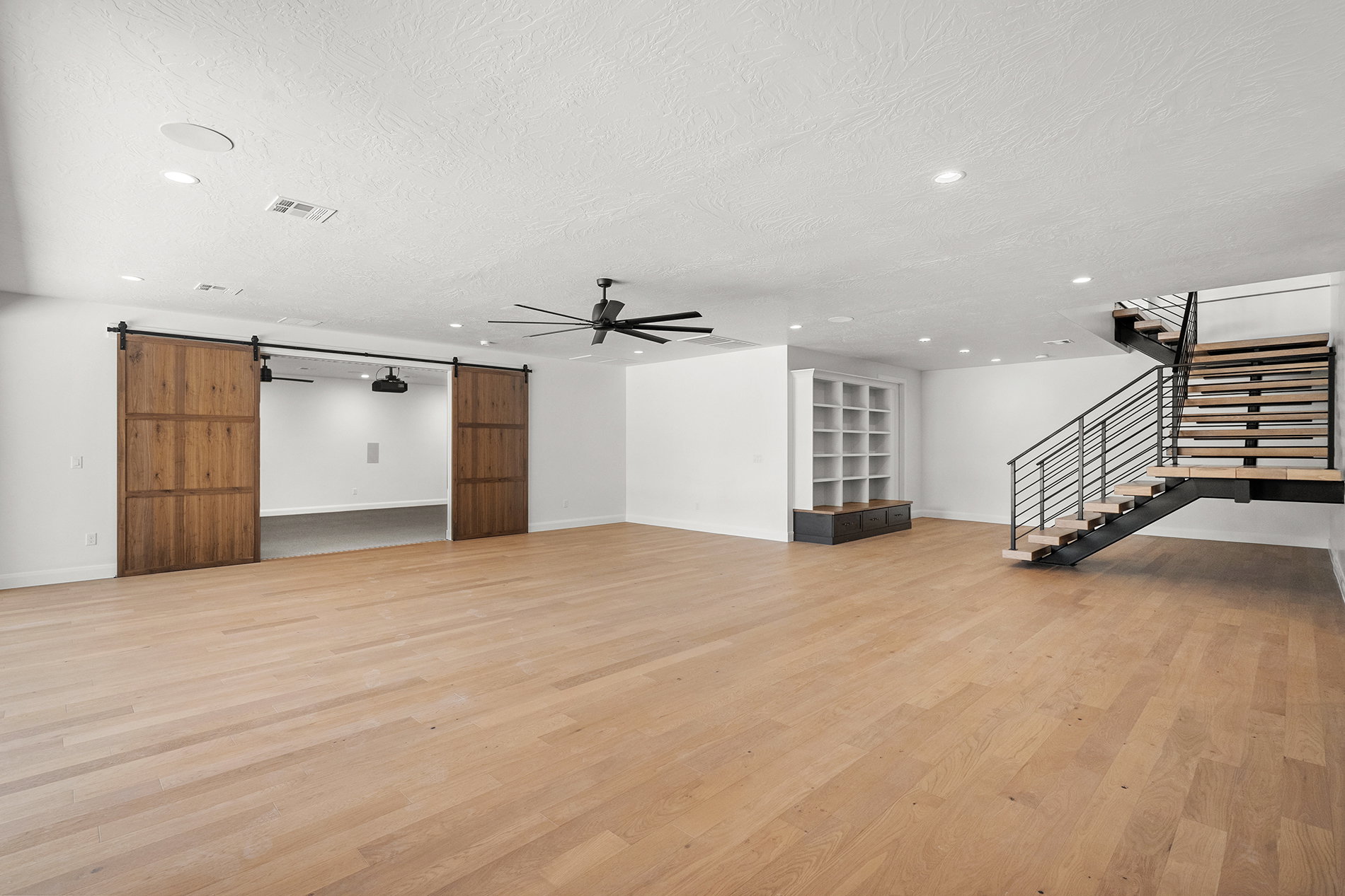 Spacious open-concept room featuring light wood-finish flooring, a modern black metal and wood staircase, and built-in shelving with a bench