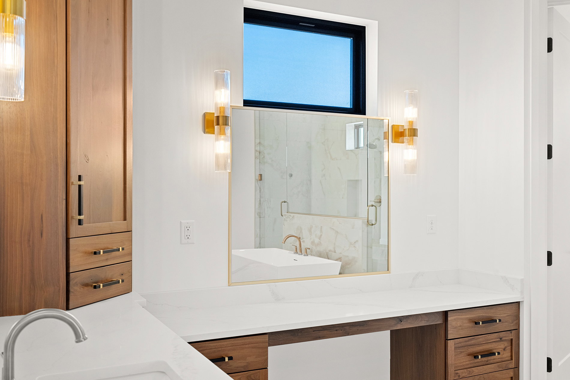 White countertop vanity with integrated sink, wood-finish cabinetry, and gold-toned wall sconces
