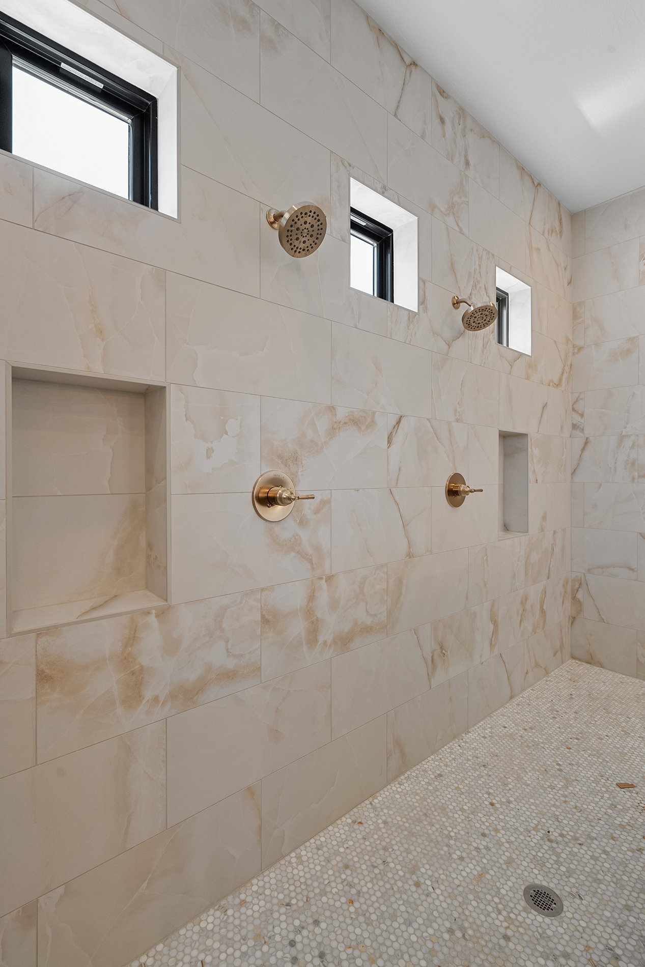 Spacious shower featuring floor-to-ceiling marble-look tile, dual rainfall showerheads, two integrated wall niches, and a mosaic pebble floor