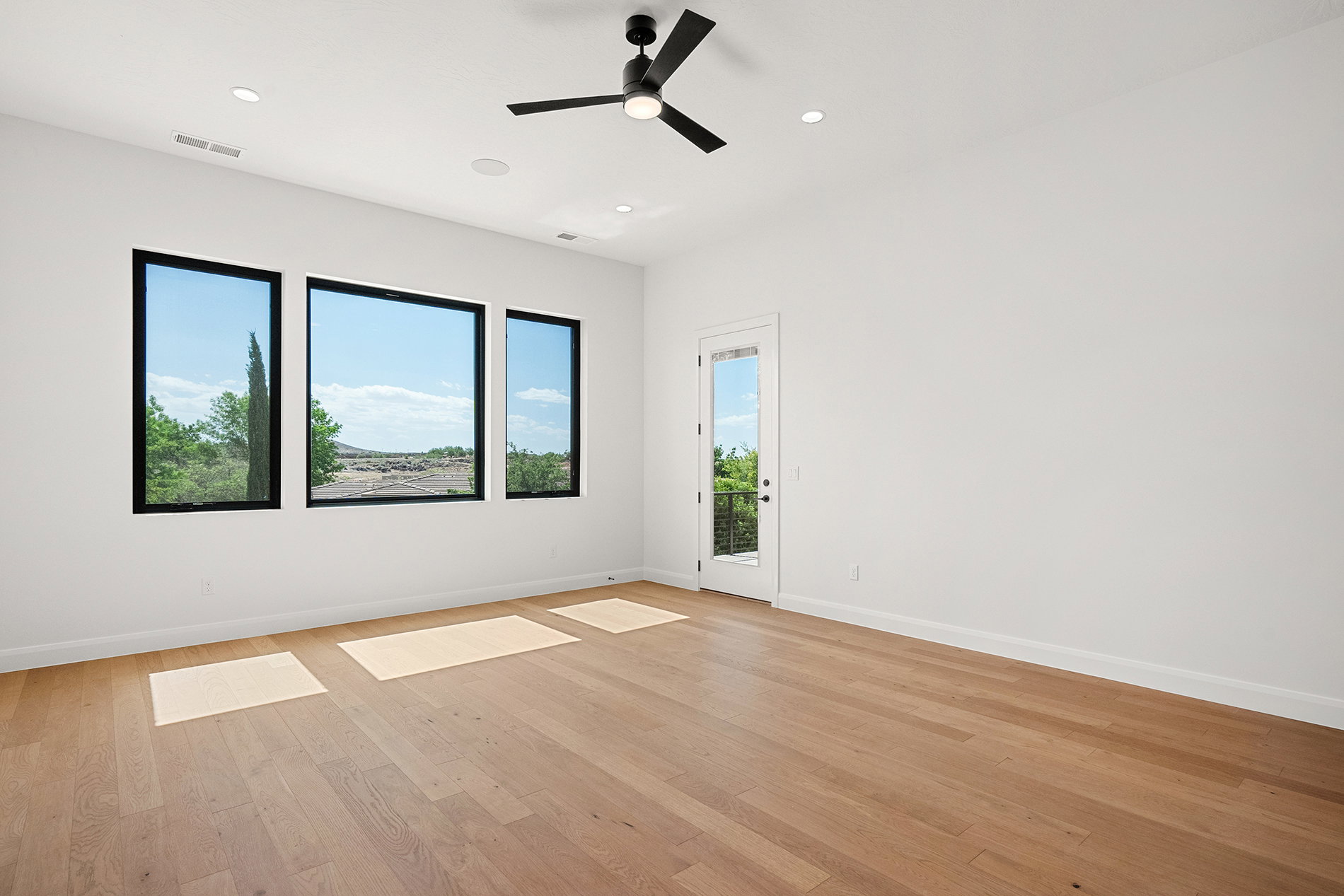 Expansive room featuring light wood-finish flooring, three large rectangular windows with black frames, and a glass-paneled door with a black railing