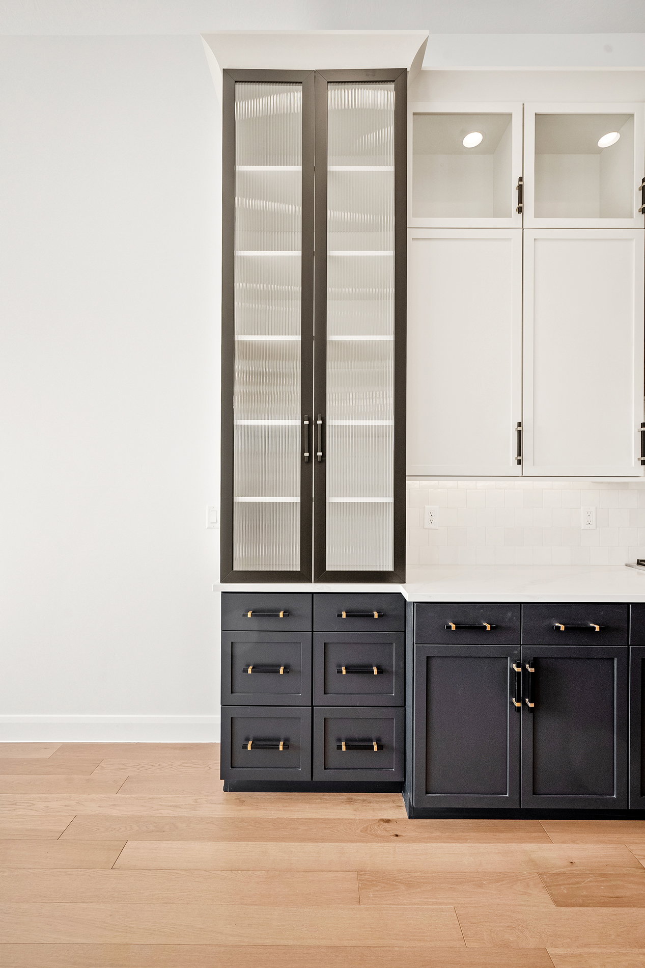 Kitchen cabinetry featuring a tall pantry with reeded glass doors, contrasting white upper cabinets with integrated lighting, dark lower cabinets, and brass hardware