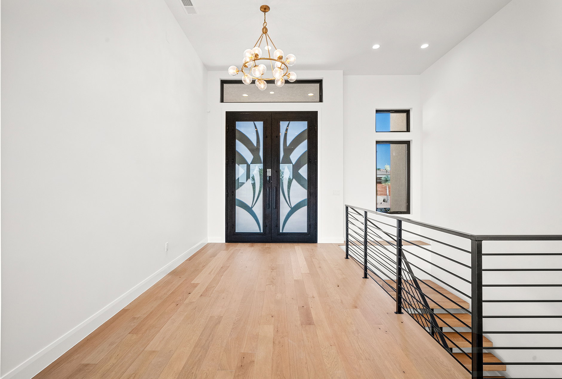 Double entry doors with intricate glass detailing and a transom window, complemented by a contemporary chandelier, hardwood flooring, and a horizontal metal railing