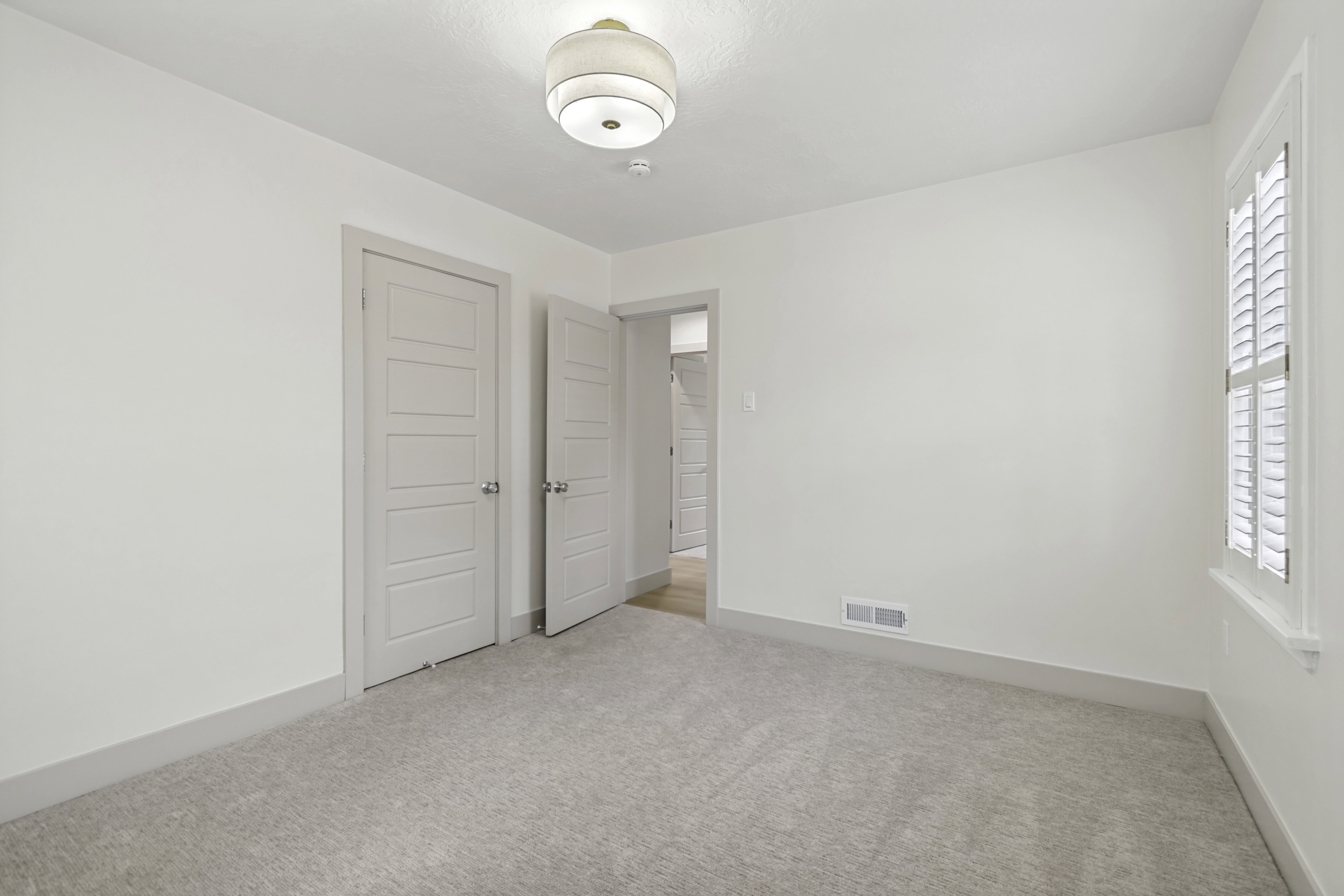 Carpeted room featuring a ceiling-mounted light fixture, white walls, and a window with plantation shutters
