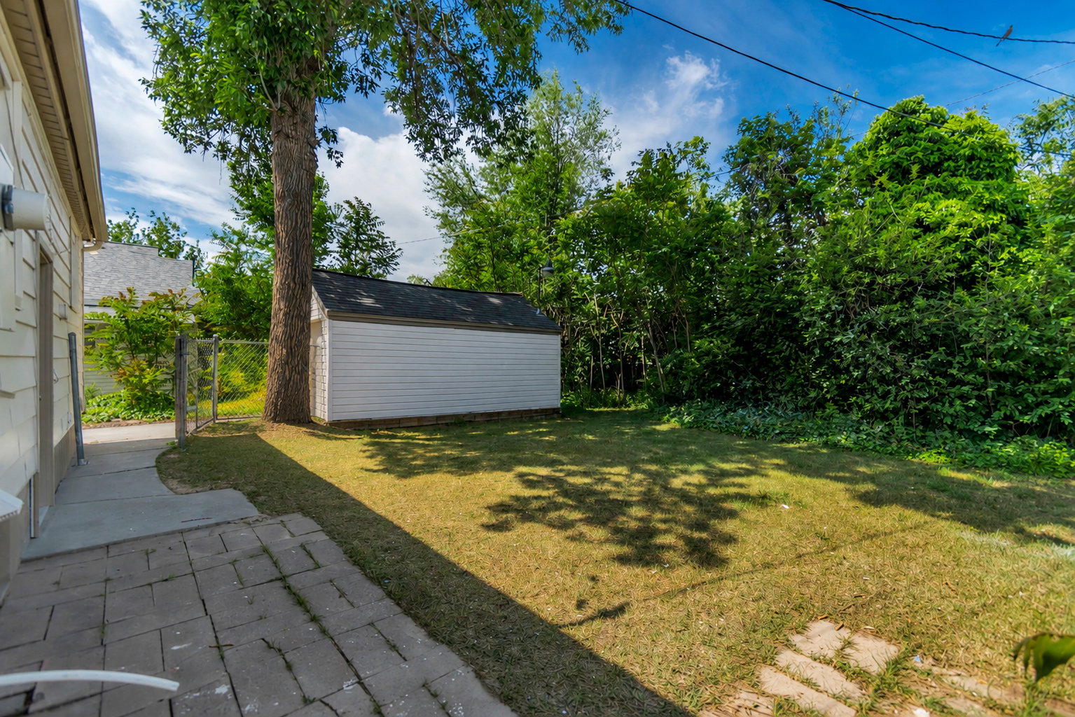 Expansive backyard featuring a white storage shed with dark roofing, a mature tree providing natural shade, a partial chain-link fence, and a stone paver patio