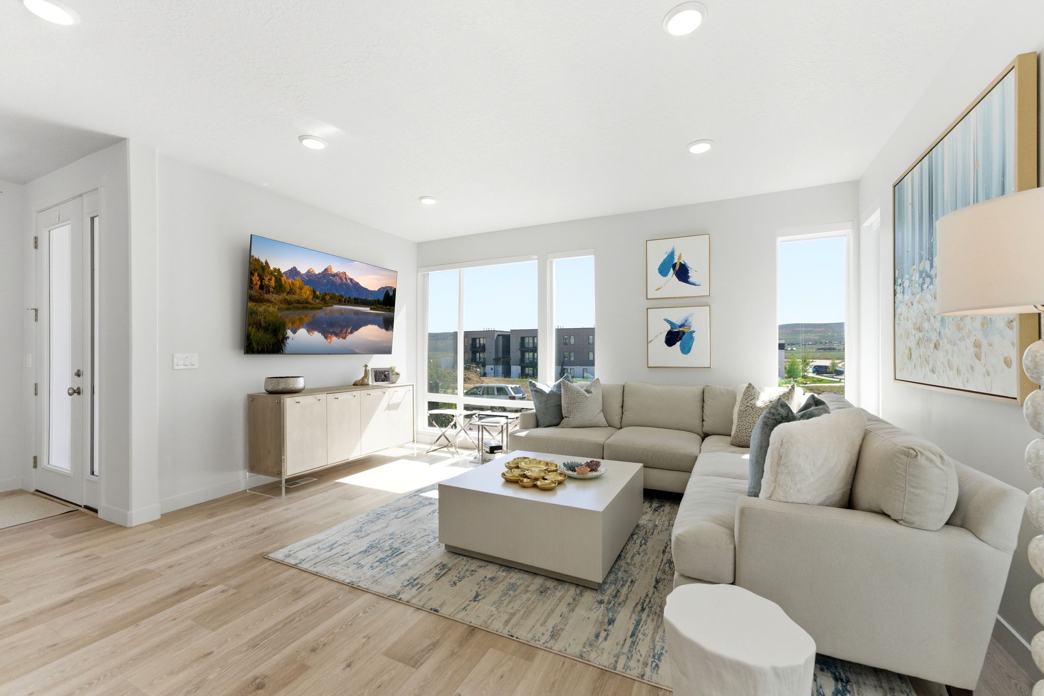 Open-concept living space with light wood-finish flooring, recessed lighting, expansive windows, a built-in wall-mounted television, and a white entry door with frosted glass paneling