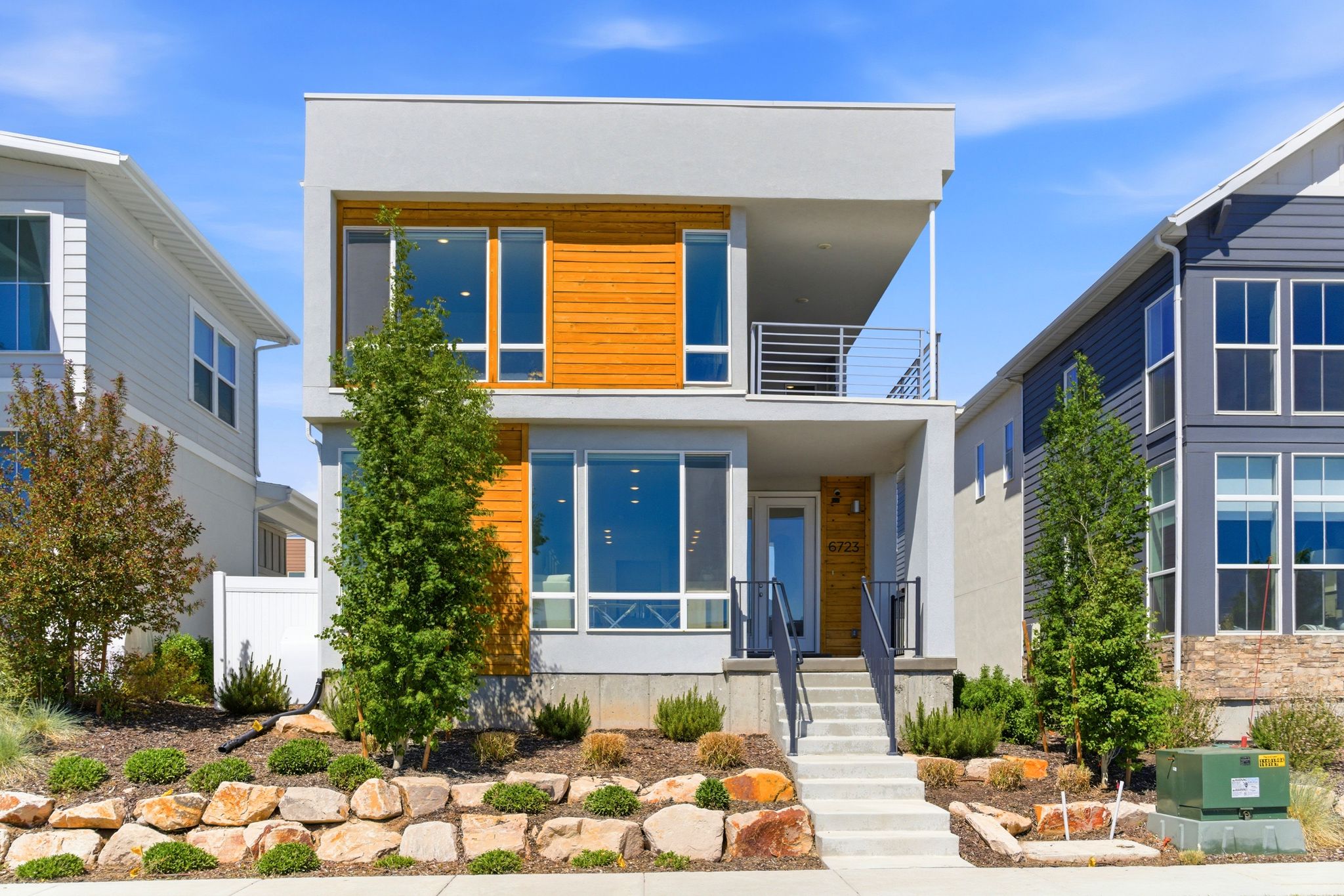 Contemporary facade featuring mixed materials, a second-story balcony with metal railing, an integrated front porch, large picture windows, and a tiered rock and plant landscape