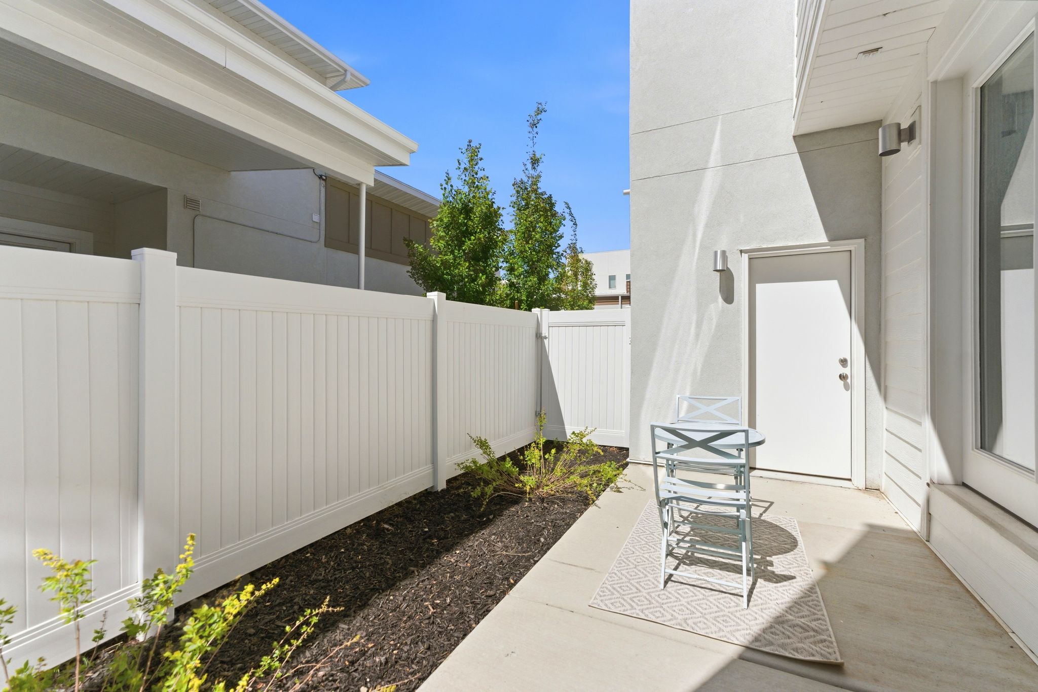 Private outdoor patio space featuring a concrete slab, white privacy fencing, exterior door access, and wall-mounted lighting fixtures